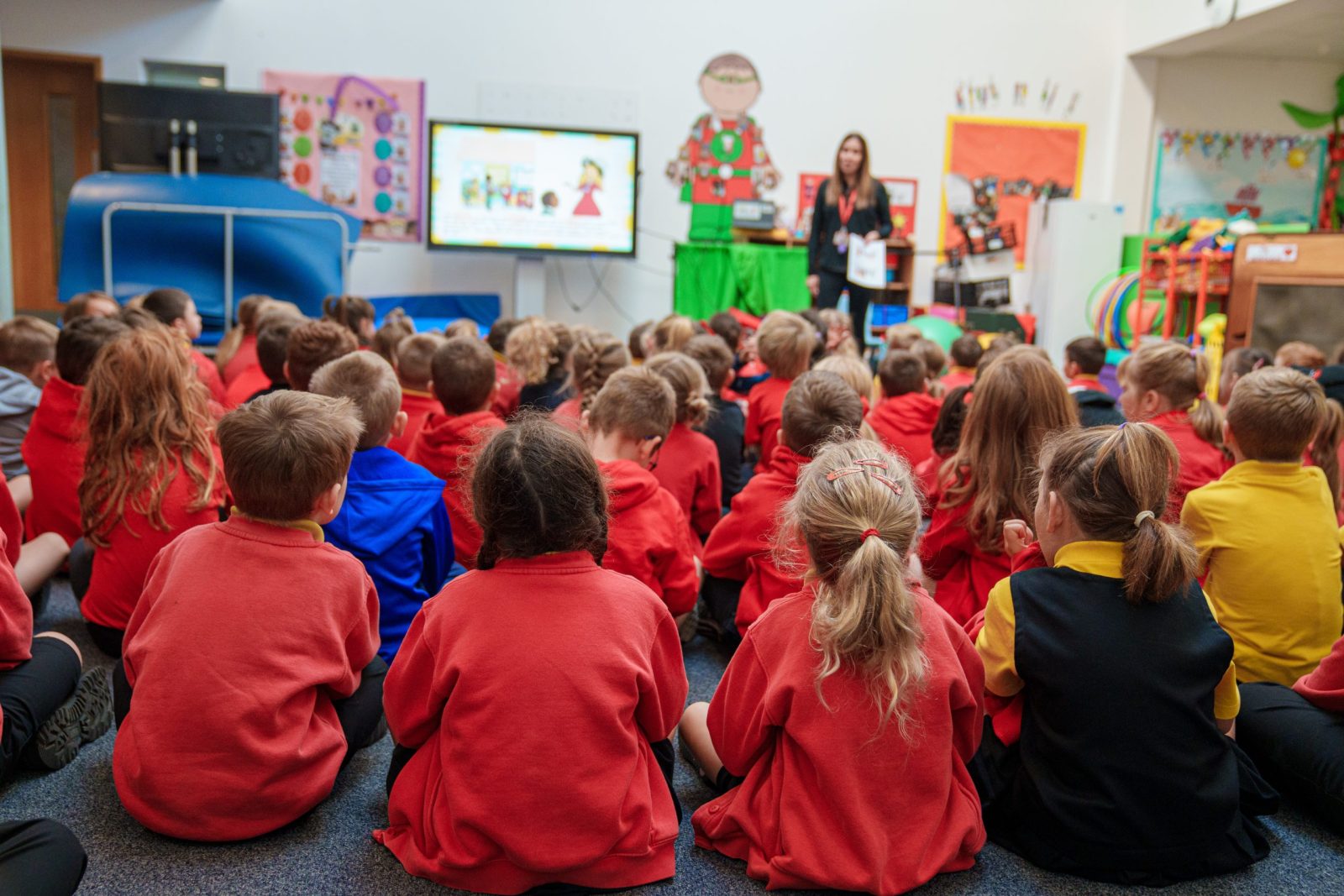 Grŵp mawr o blant ifanc mewn gwisgoedd ysgol coch yn eistedd ar y llawr, yn wynebu athro sy'n rhoi cyflwyniad mewn ystafell ddosbarth liwgar.