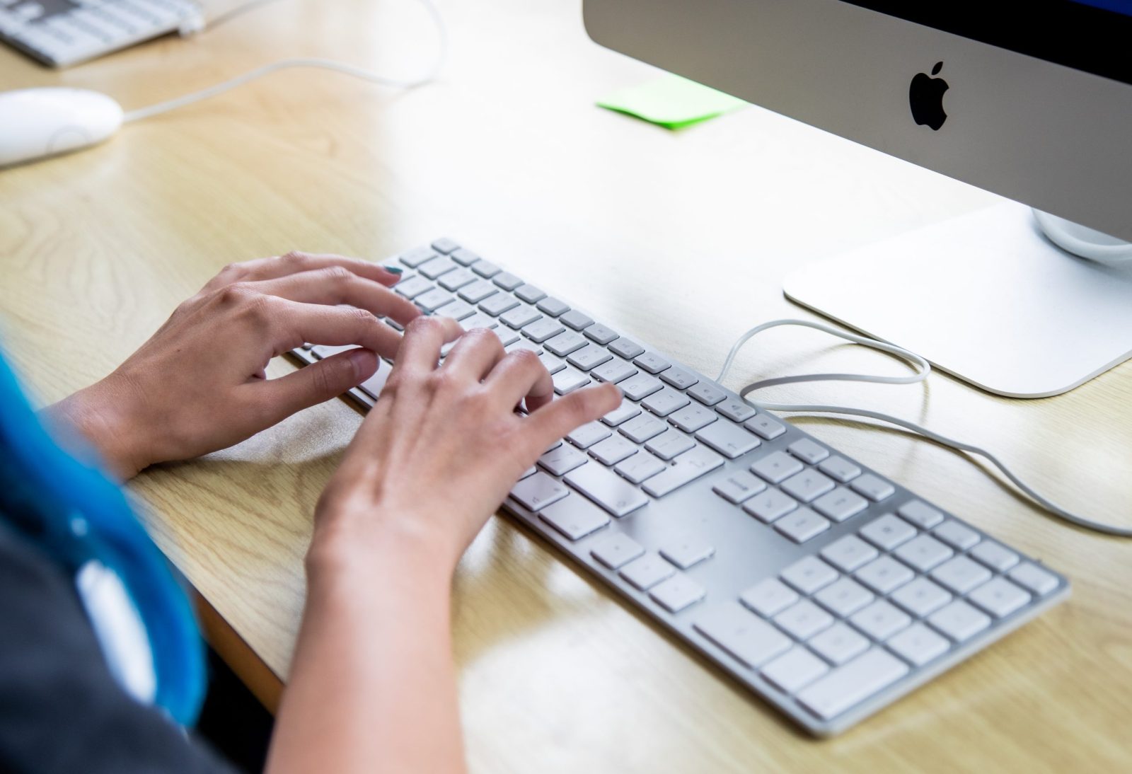 Close-up of hands typing on an Apple keyboard connected to an iMac, possibly engaged in a task involving computer work.