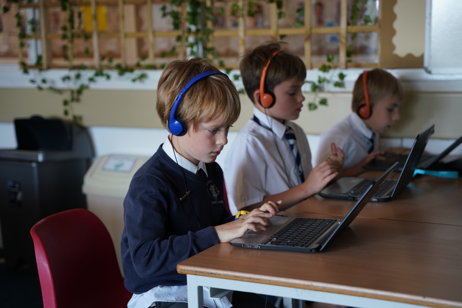 Three students in school uniforms are sitting at desks, working on laptops with headphones on, focusing on their tasks.