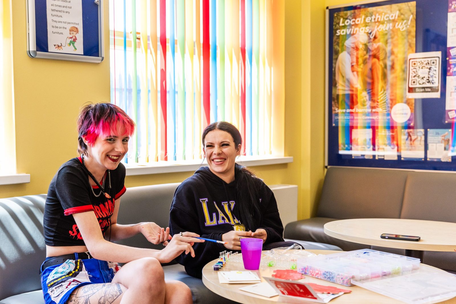 Two young women with colorful hair and casual clothing are sitting on a couch, laughing together. They are working on a craft project, with a cup and various supplies on a table in front of them.