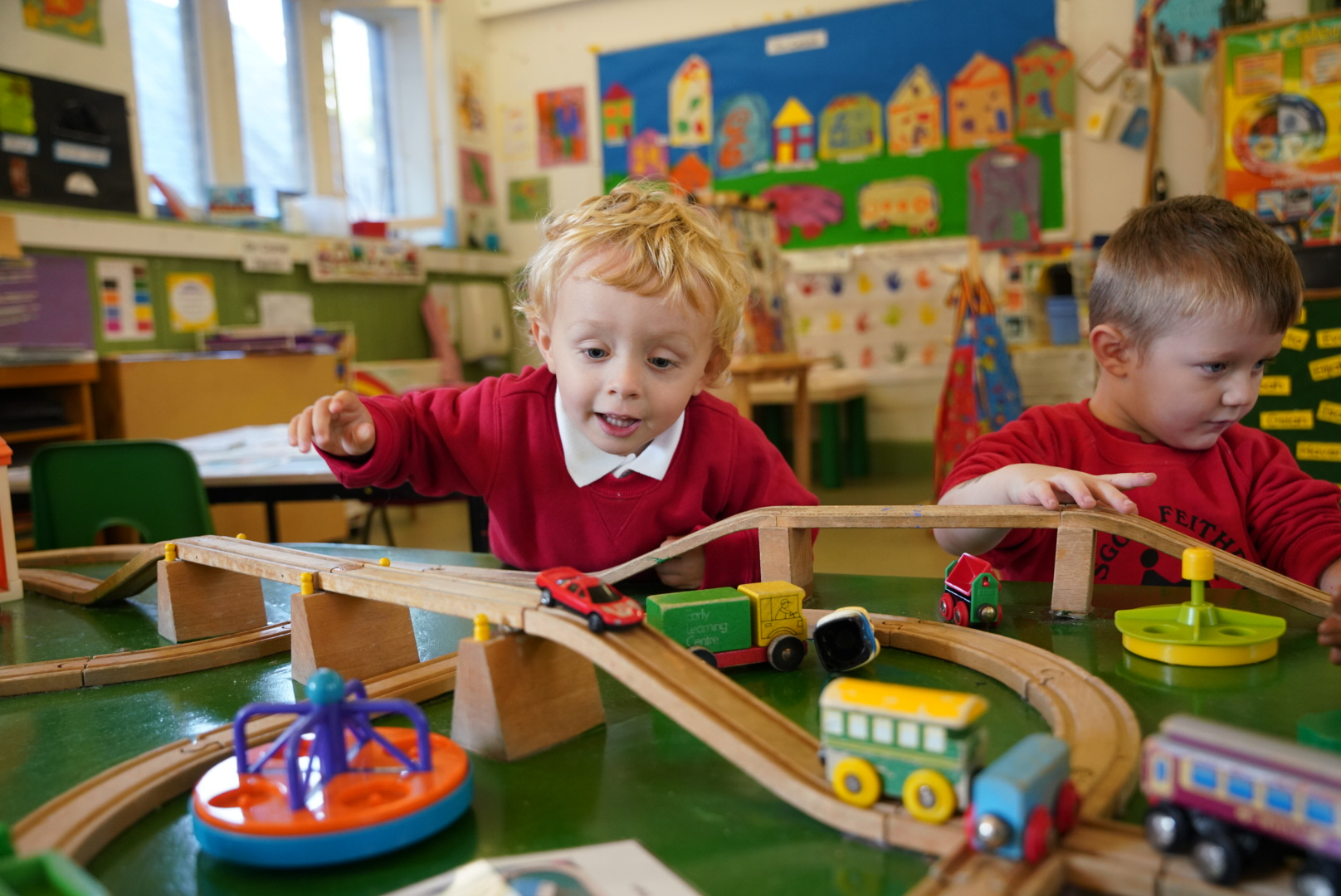 A young boy in a red school jumper playing enthusiastically with a wooden toy train set in a colorful classroom.