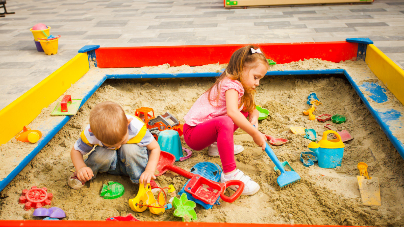 Two children playing in a sandbox with various colorful toys.