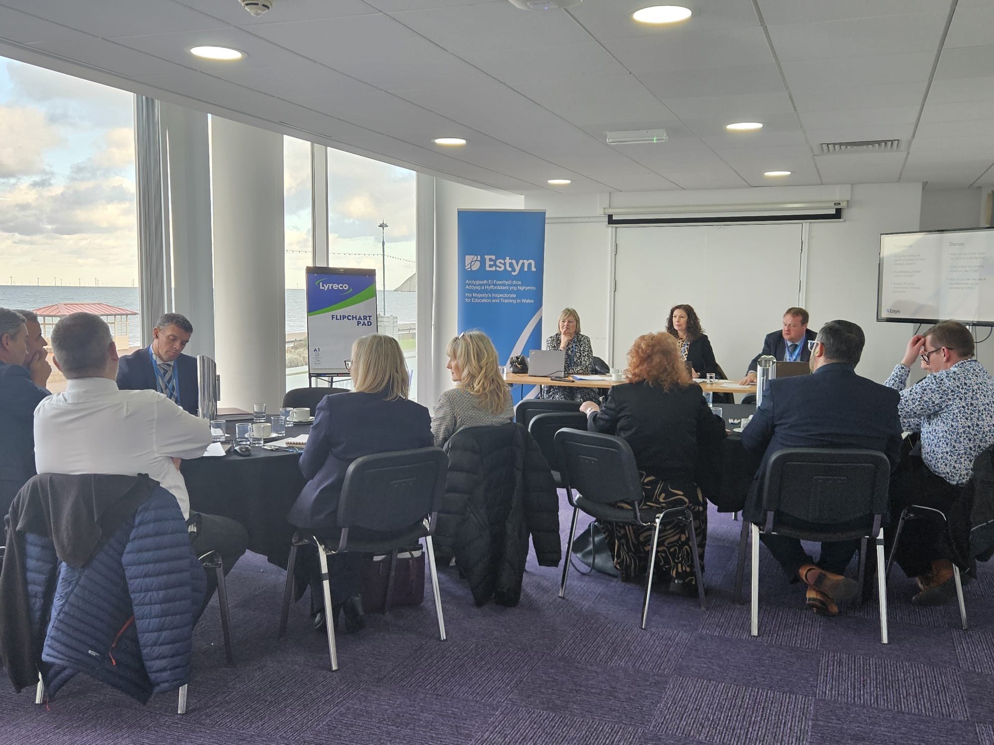 Business professionals sitting around a conference table during a meeting, with banners of Estyn visible in the background.