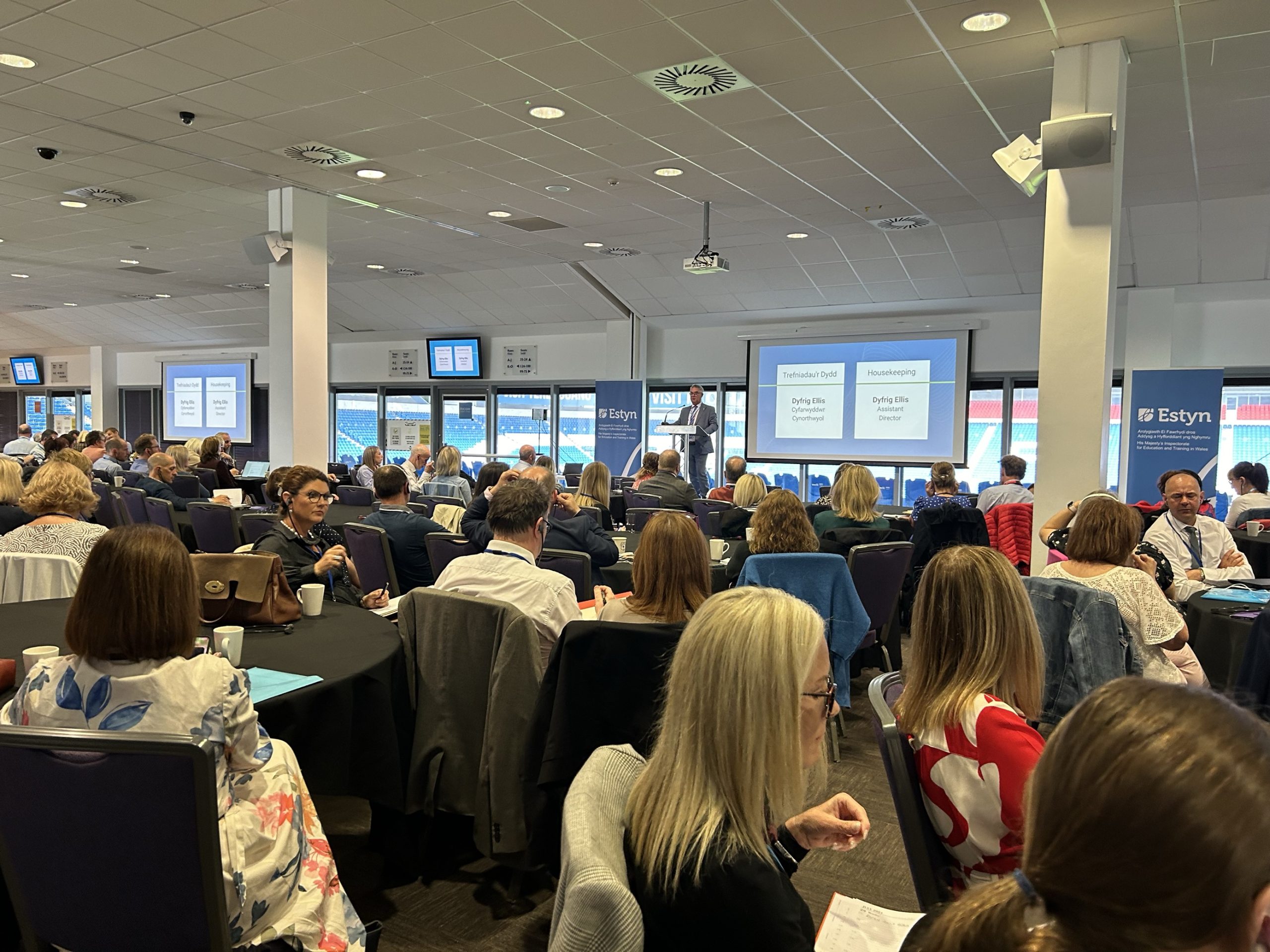 Attendees seated at a conference within a room displaying 'Estyn' banners, listening to a speaker presenting on a stage with a projected screen.